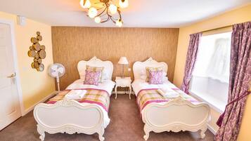 A hotel room with two ornate beds, a wooden accent wall, a chandelier, and a window with patterned curtains.