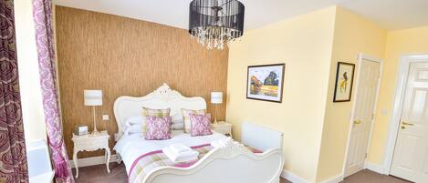 A bedroom with a white ornate bed, a wooden headboard, a chandelier, a nightstand, and a door.