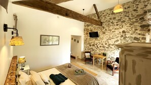 A bedroom with a bed, a wooden headboard, a stone wall, a television, and a dining area with a table and chairs. - La Petite Félixière, B&B indépendants à 10 min de Cholet, 25 min du Puy du Fou et 35 min de Nantes (Sèvremoine)