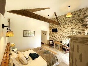 A bedroom with a bed, a wooden headboard, a stone wall, a television, and a dining area with a table and chairs.