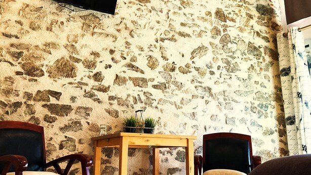 A room with a stone wall, a flat-screen TV mounted on it, a wooden table, two chairs, and a hanging light fixture. - La Petite Félixière, B&B indépendants à 10 min de Cholet, 25 min du Puy du Fou et 35 min de Nantes (Sèvremoine)