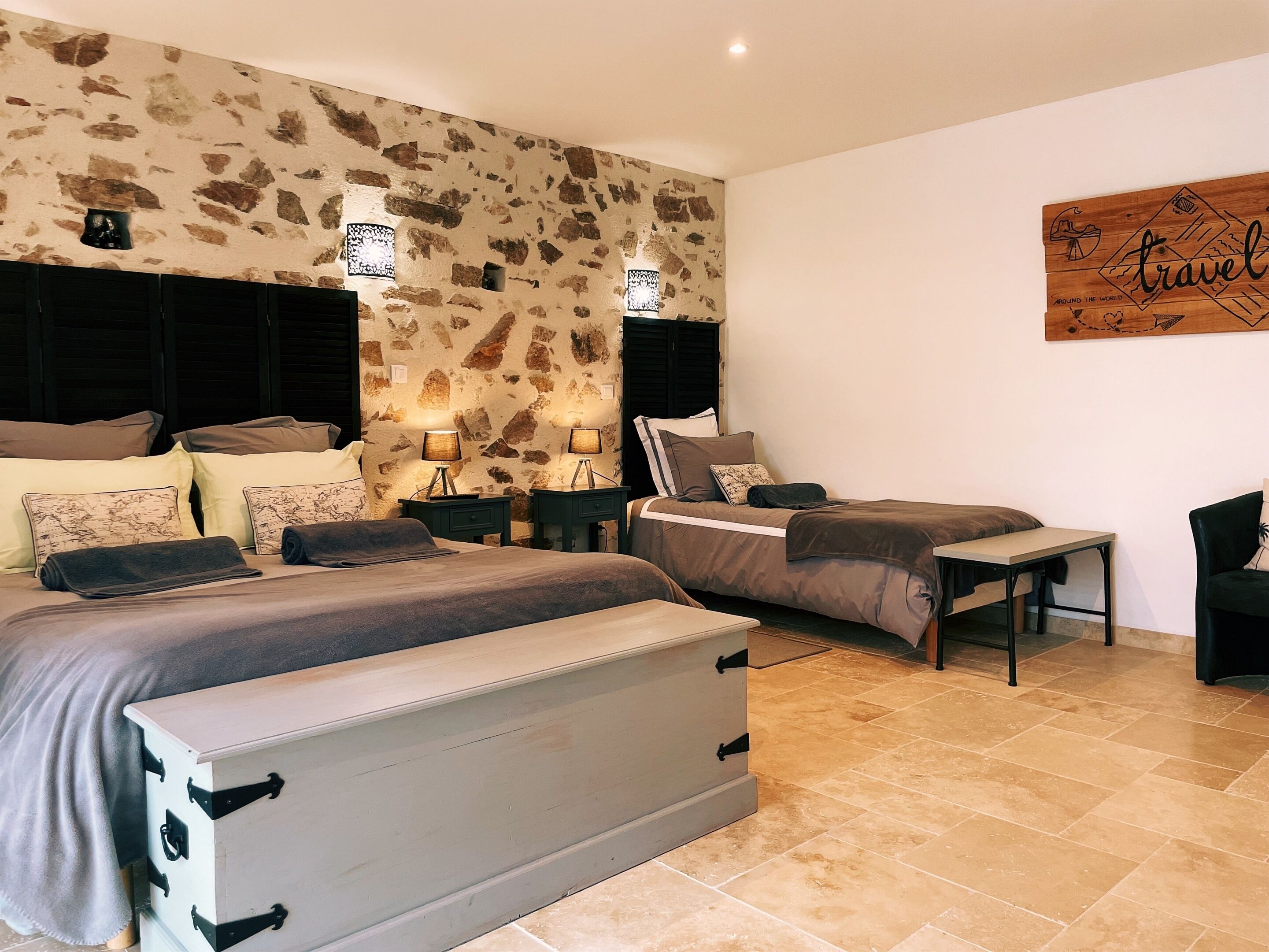 A bedroom with a stone wall, two beds, bedside tables with lamps, a bench, and a travel-themed artwork.
