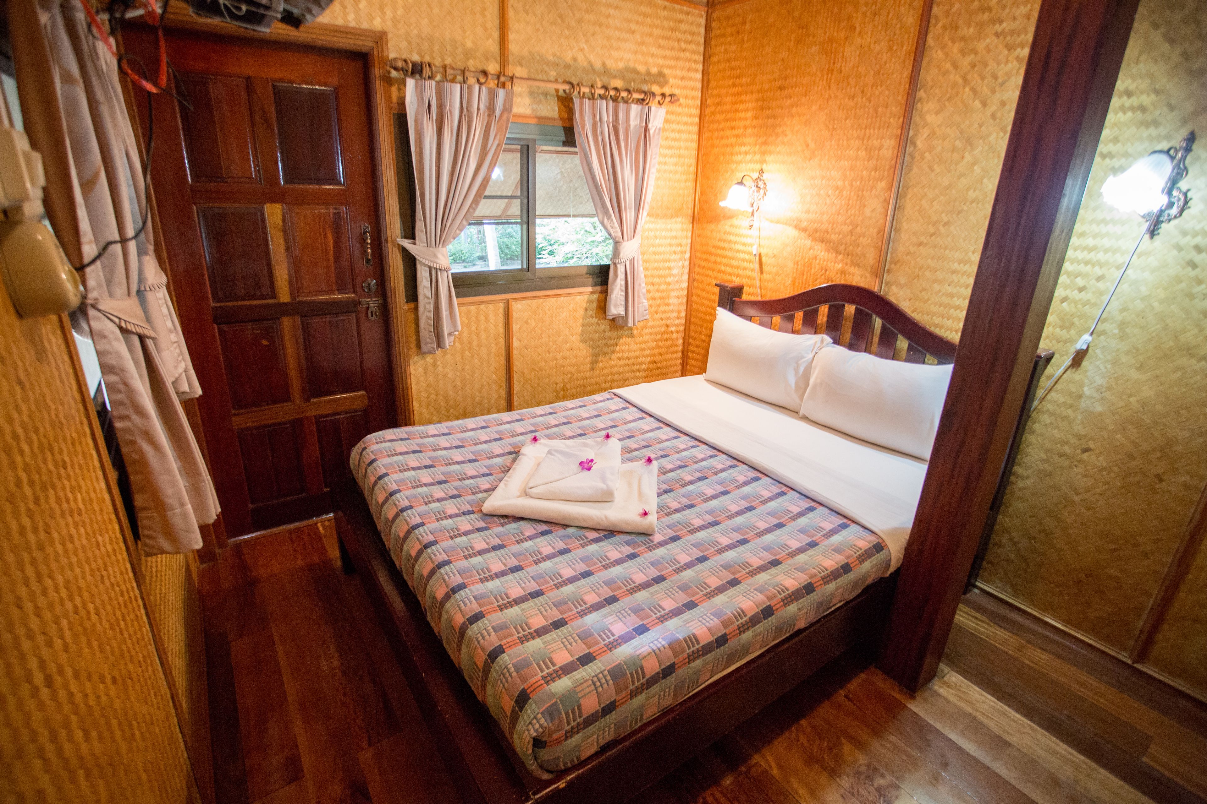 Un dormitorio con cama de madera, ropa de cama escocesa, una ventana con cortinas y una lámpara fijada a la pared.