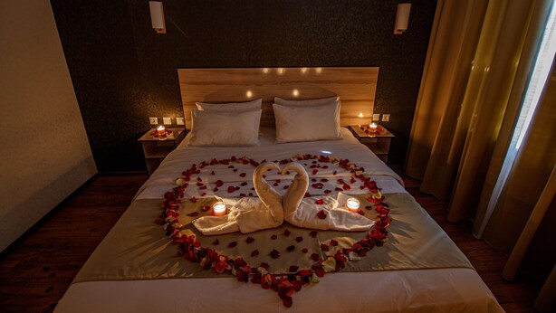 A hotel room with a bed adorned with rose petals and two swan-shaped towels, lit by bedside lamps. - Level Hotel (Osh)