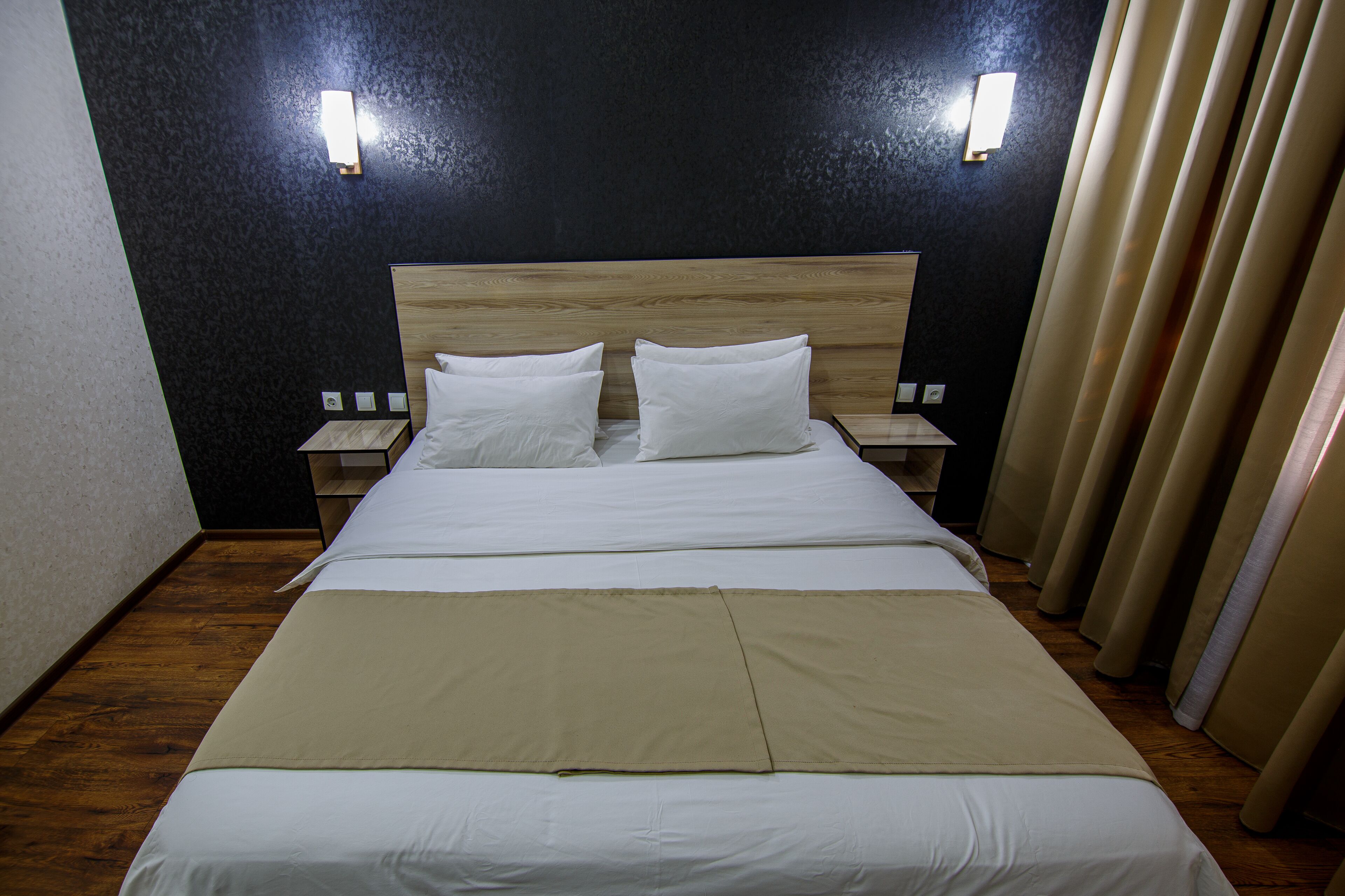 Una cama bien hecha con sábanas blancas y dos almohadas, cabecera de madera, mesitas de noche con lámparas y una pared oscura con dos luces empotradas.
