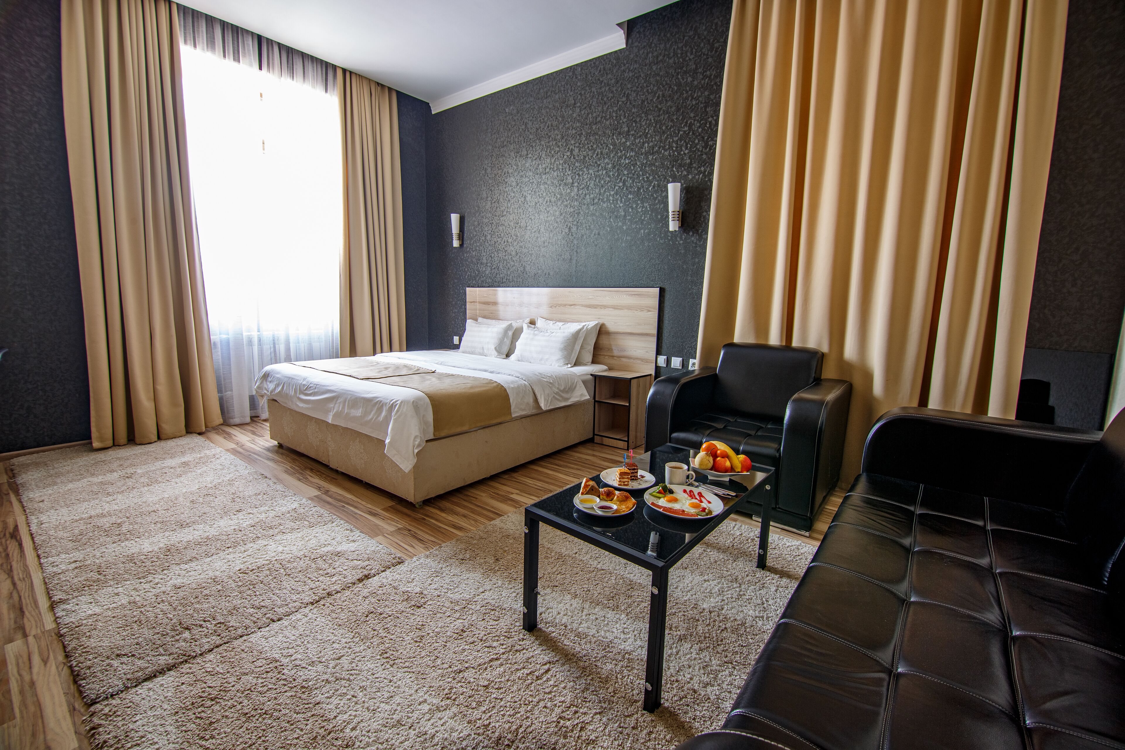 A hotel room with a bed, a black leather sofa, a small table with breakfast items, and a large window with curtains.
