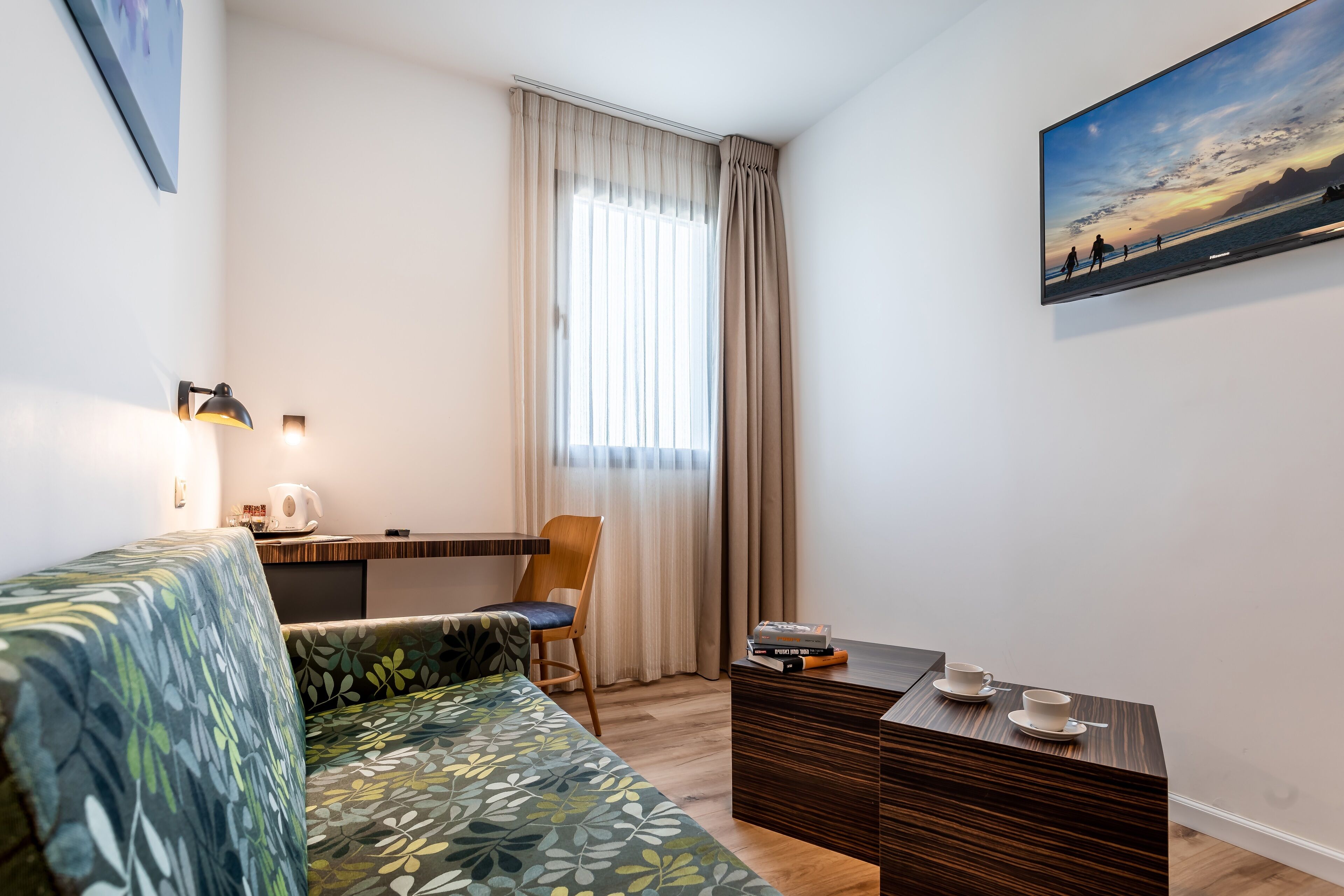 A modern hotel room with a sofa, a small table with cups, a chair, a desk, and a painting on the wall.