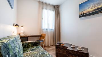 Quarto de hotel moderno com sofá, uma pequena mesa com xícaras, uma cadeira, escrivaninha e uma pintura na parede.