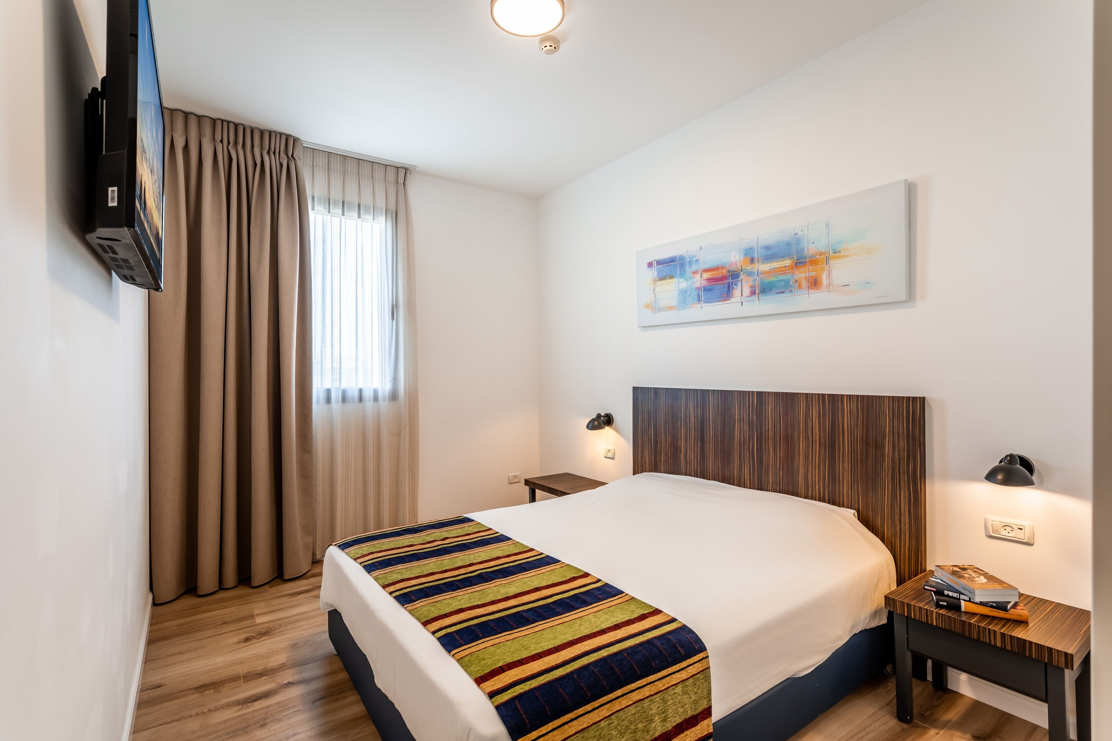 A hotel room with a large bed, a flat-screen TV mounted on the wall, a bedside table with a lamp, and a window with curtains.