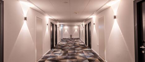A hotel hallway with patterned carpet, wall-mounted lights, and doors on both sides.