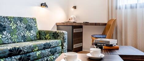 A modern hotel room with a sofa, a wooden chair, a small table with two cups and saucers, a coffee maker, and a stack of books.