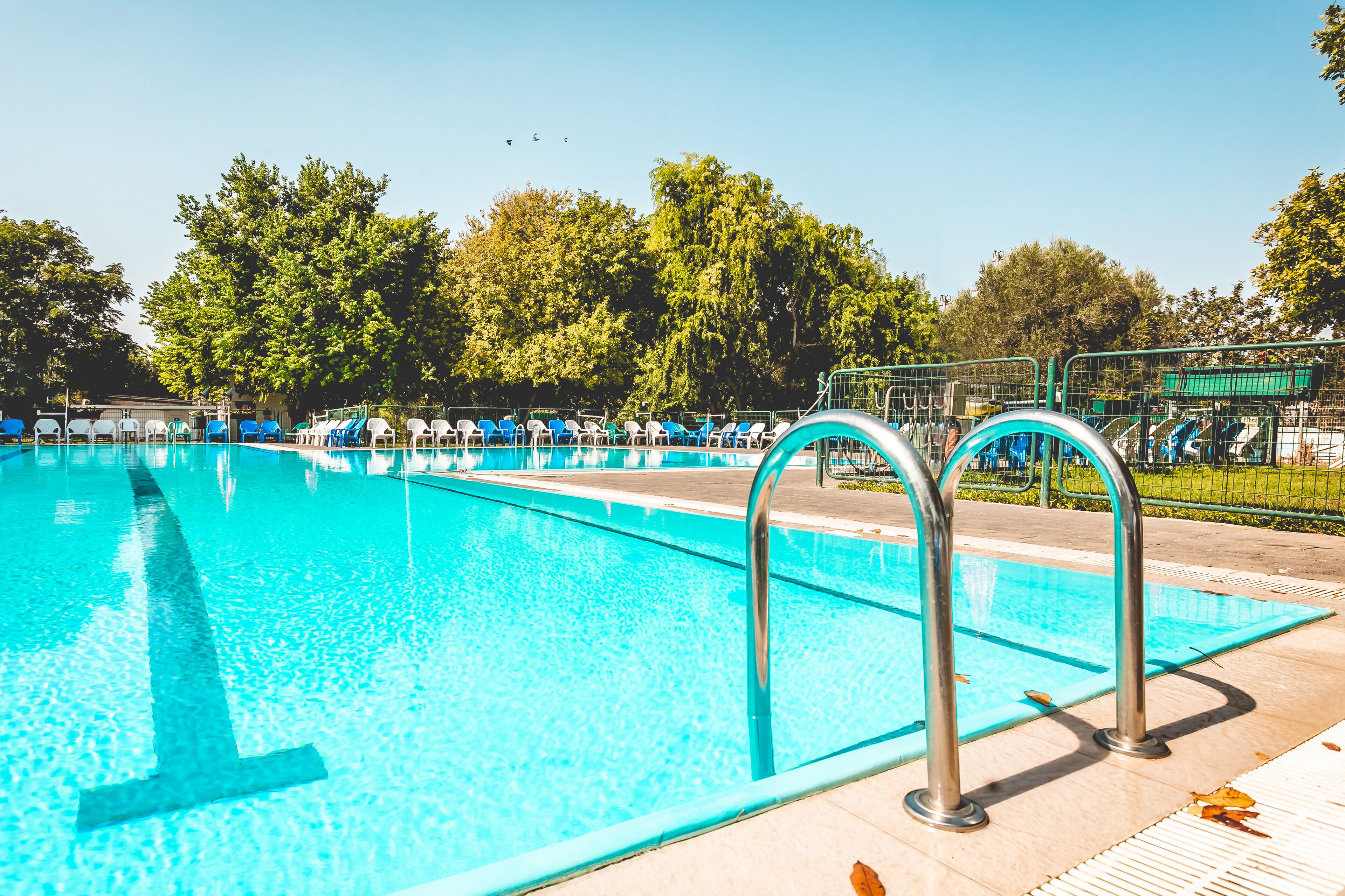 Una piscina con agua cristalina azul, rodeada por un patio de baldosas y barandillas de metal.