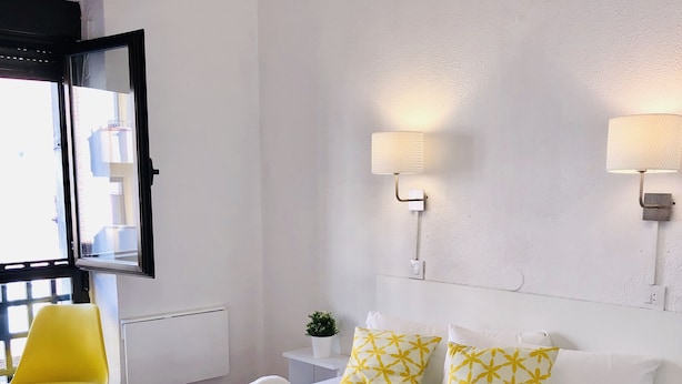 A hotel room with a large bed, a yellow chair, a wall-mounted lamp, and a window with a view of buildings. - CH Arriaza II (Madrid)