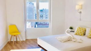 A modern bedroom with a large bed, a yellow chair, a window with a view of buildings, and a hanging light fixture. - CH Arriaza II (Madrid)