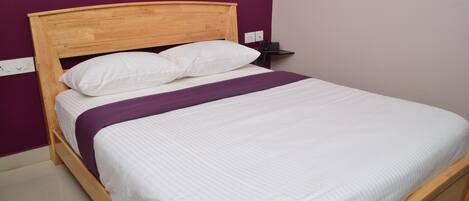 Una cama individual con cabecera de madera, ropa de cama blanca y una manta morada.