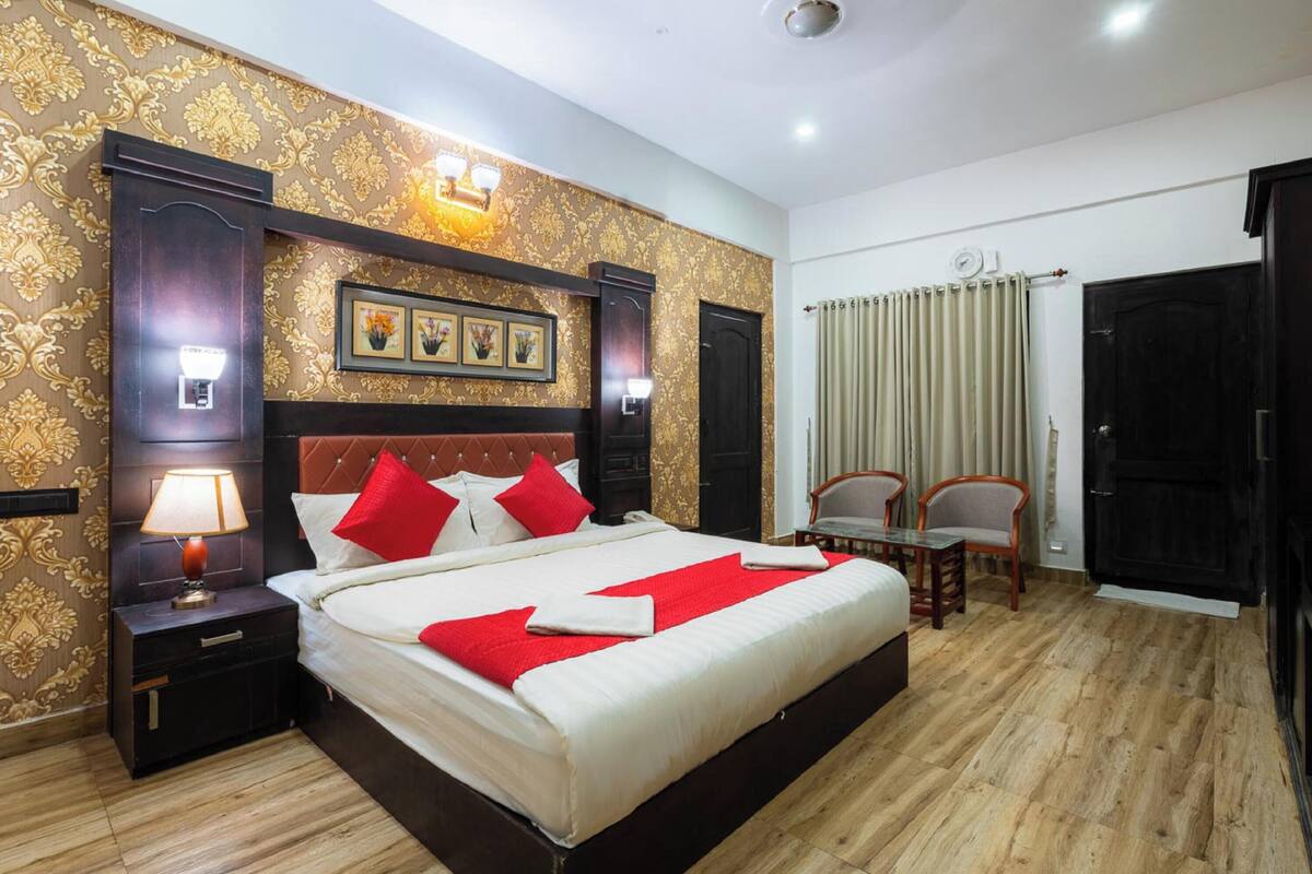 A hotel room with a large bed, wooden headboard, two bedside tables with lamps, a nightstand, a wooden floor, a window with curtains, and a door.