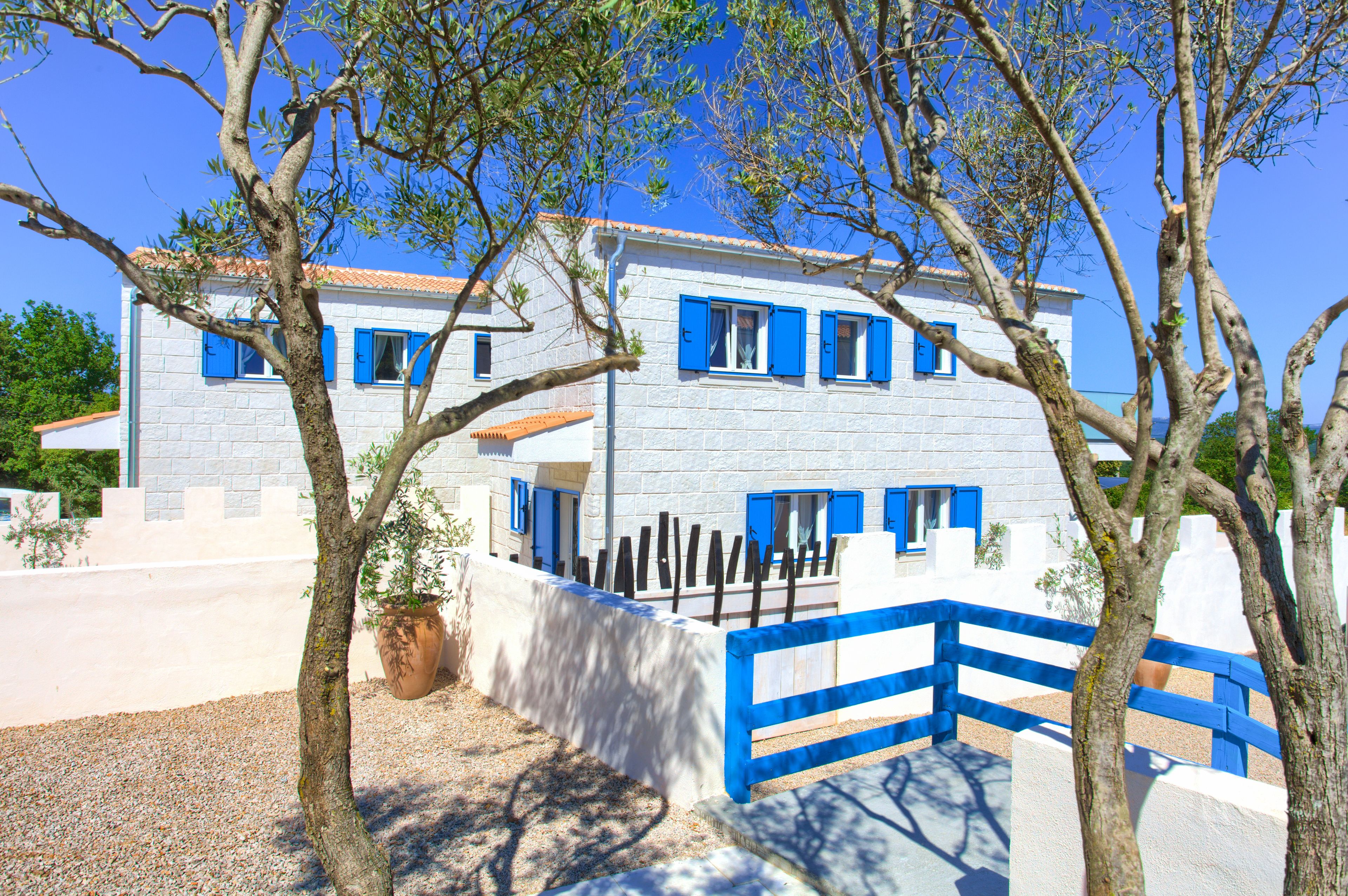 A two-story building with blue shutters and white walls, surrounded by trees and a blue fence.