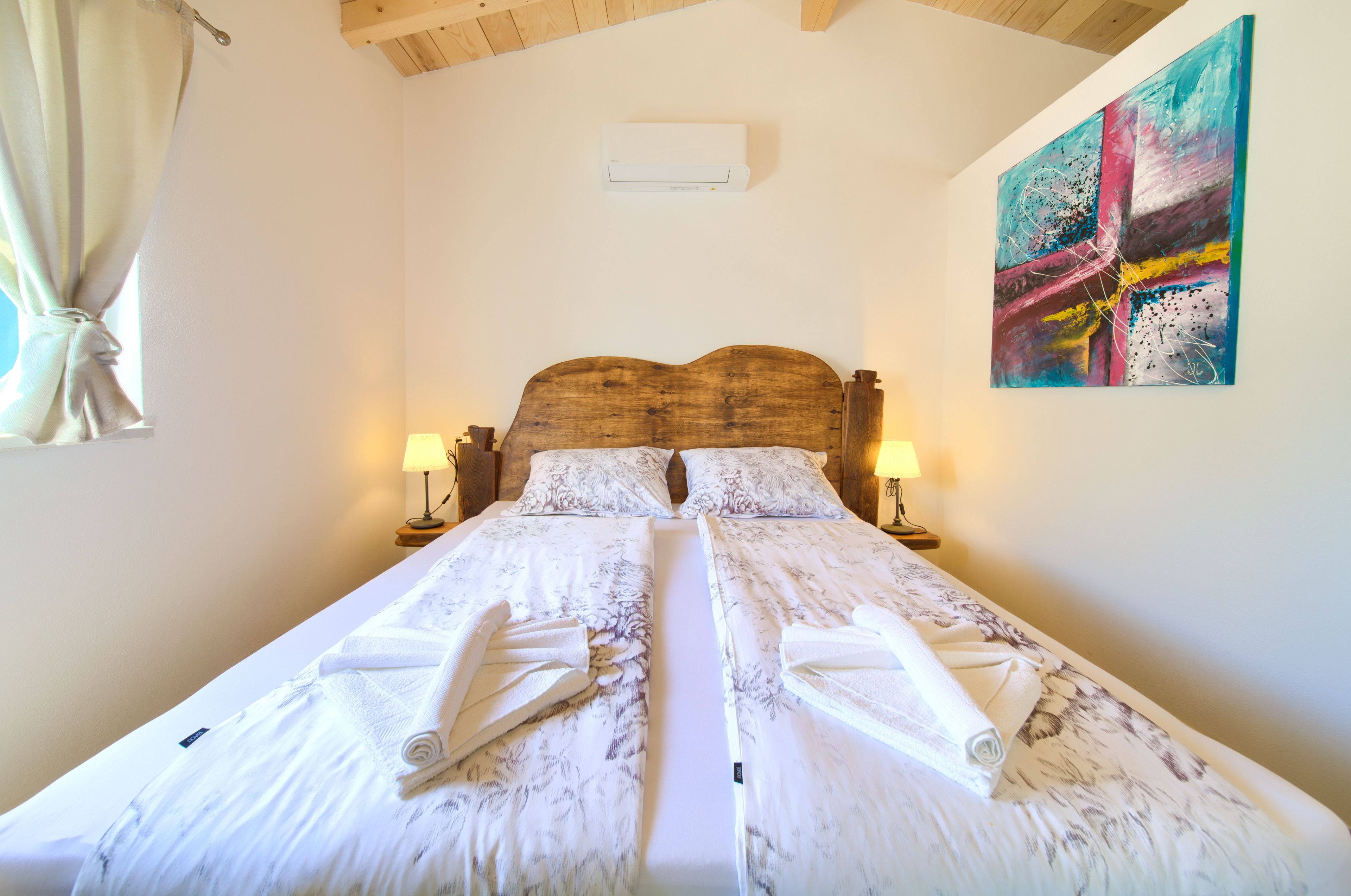 A double bed with white linens and towels, wooden headboard, bedside lamps, and a colorful abstract painting on the wall.