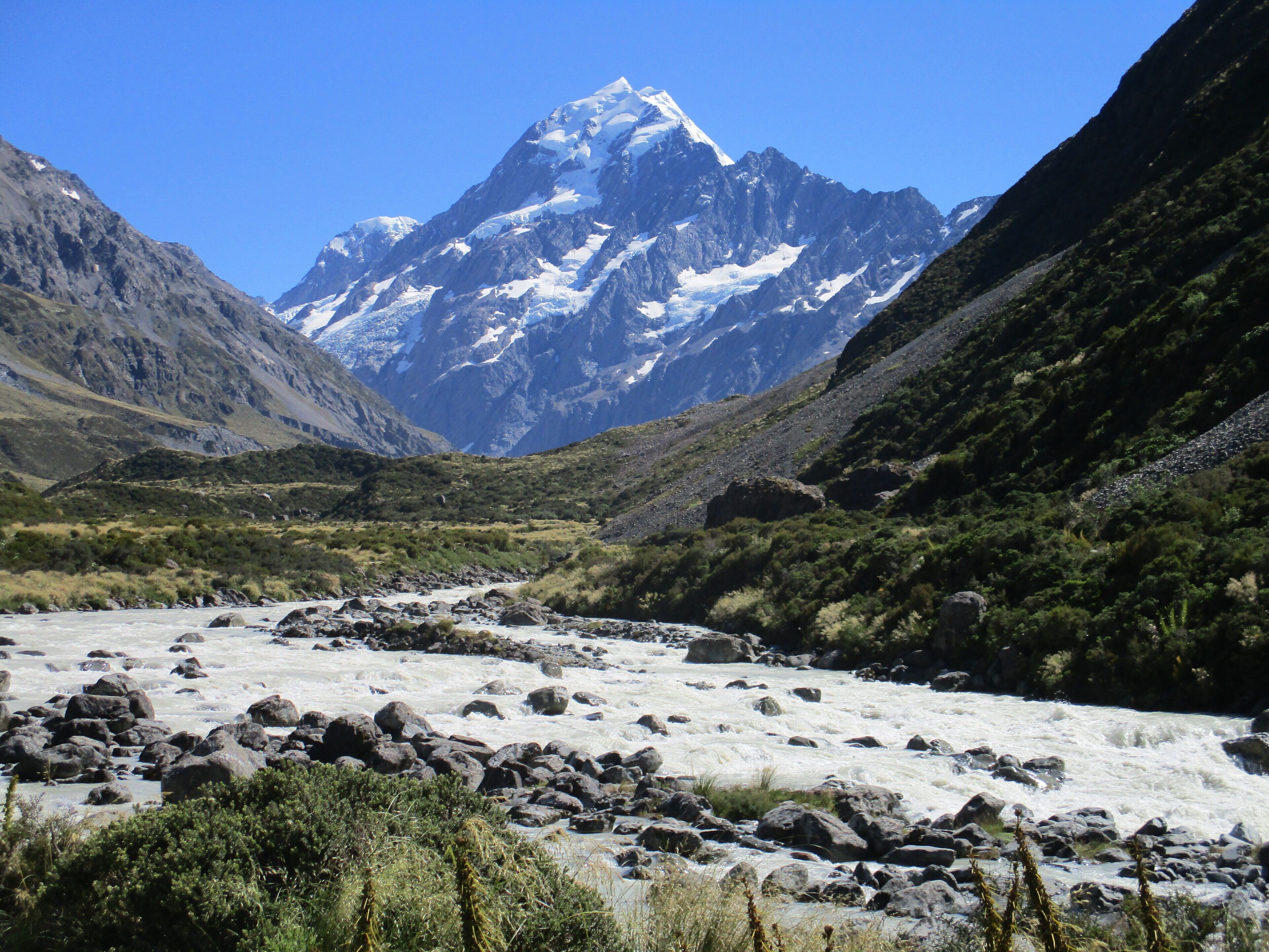 Paisagem montanhosa com pico nevado, rio com pedras e vegetação verde.