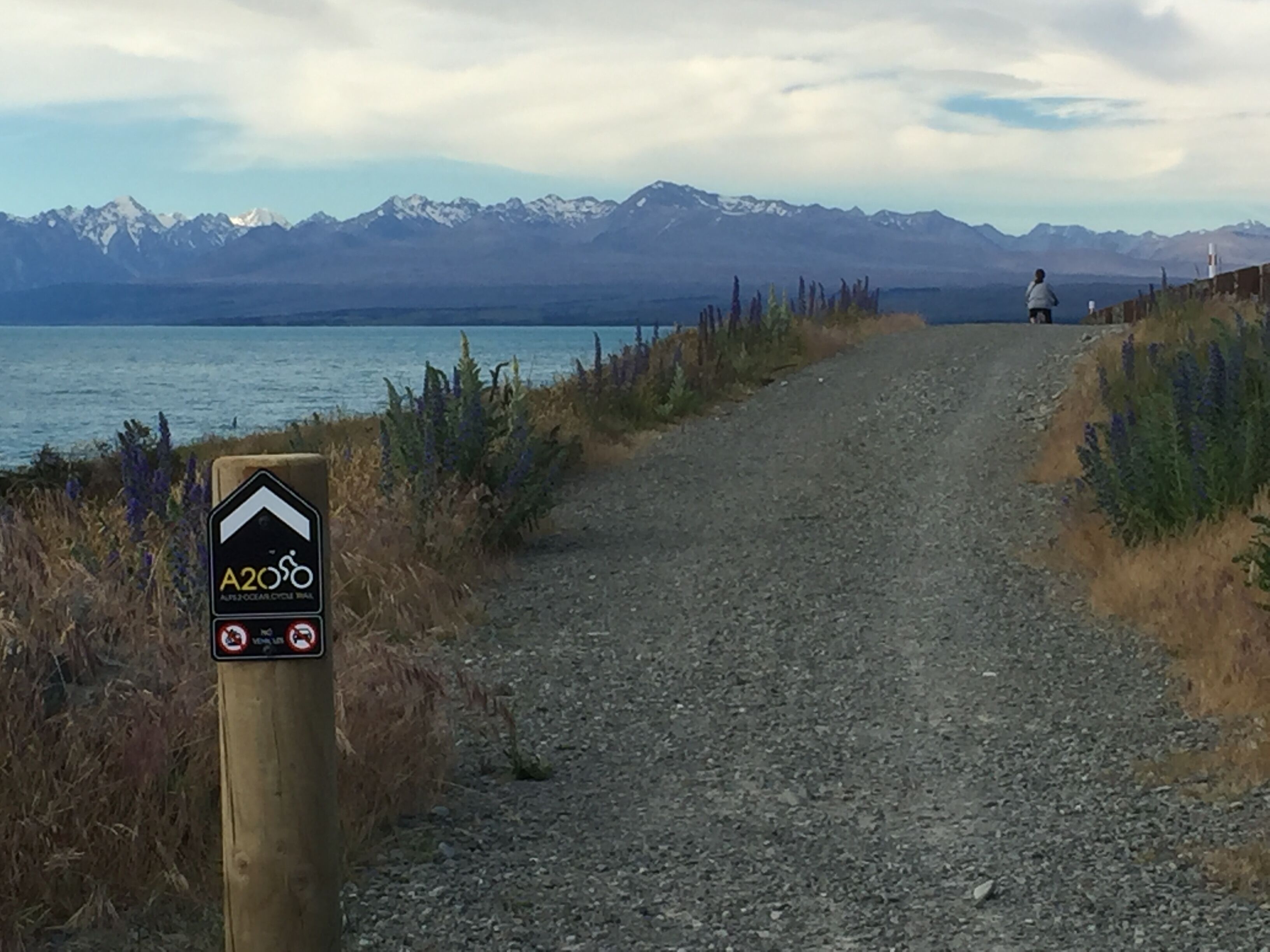 A gravel path leading to a body of water with mountains in the background and a sign indicating a cycling route.