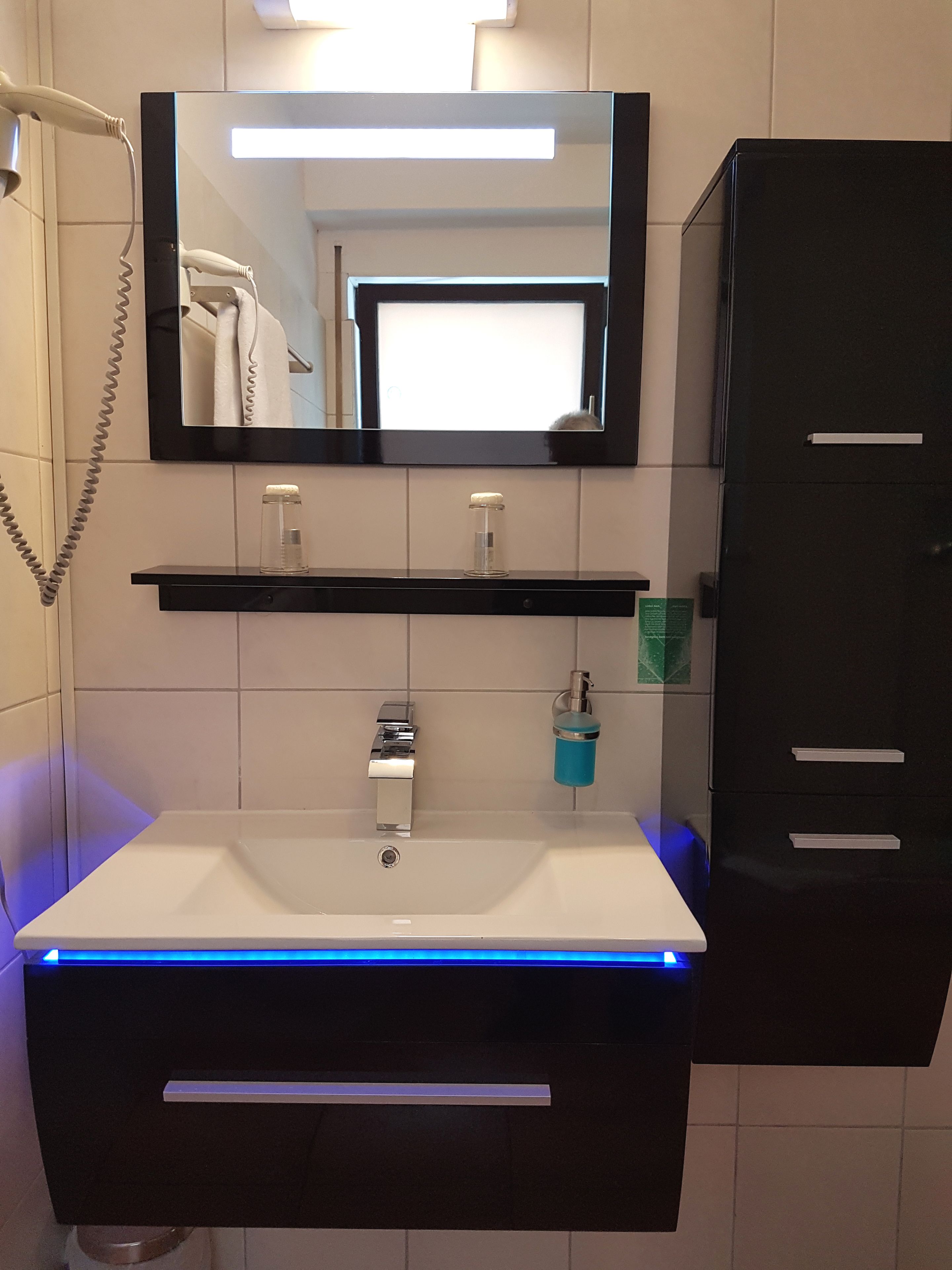 Een badkamer met een wit wastafel, een spiegel erboven, een zwart kastje rechts en een blauw verlichte wastafelmeubel.