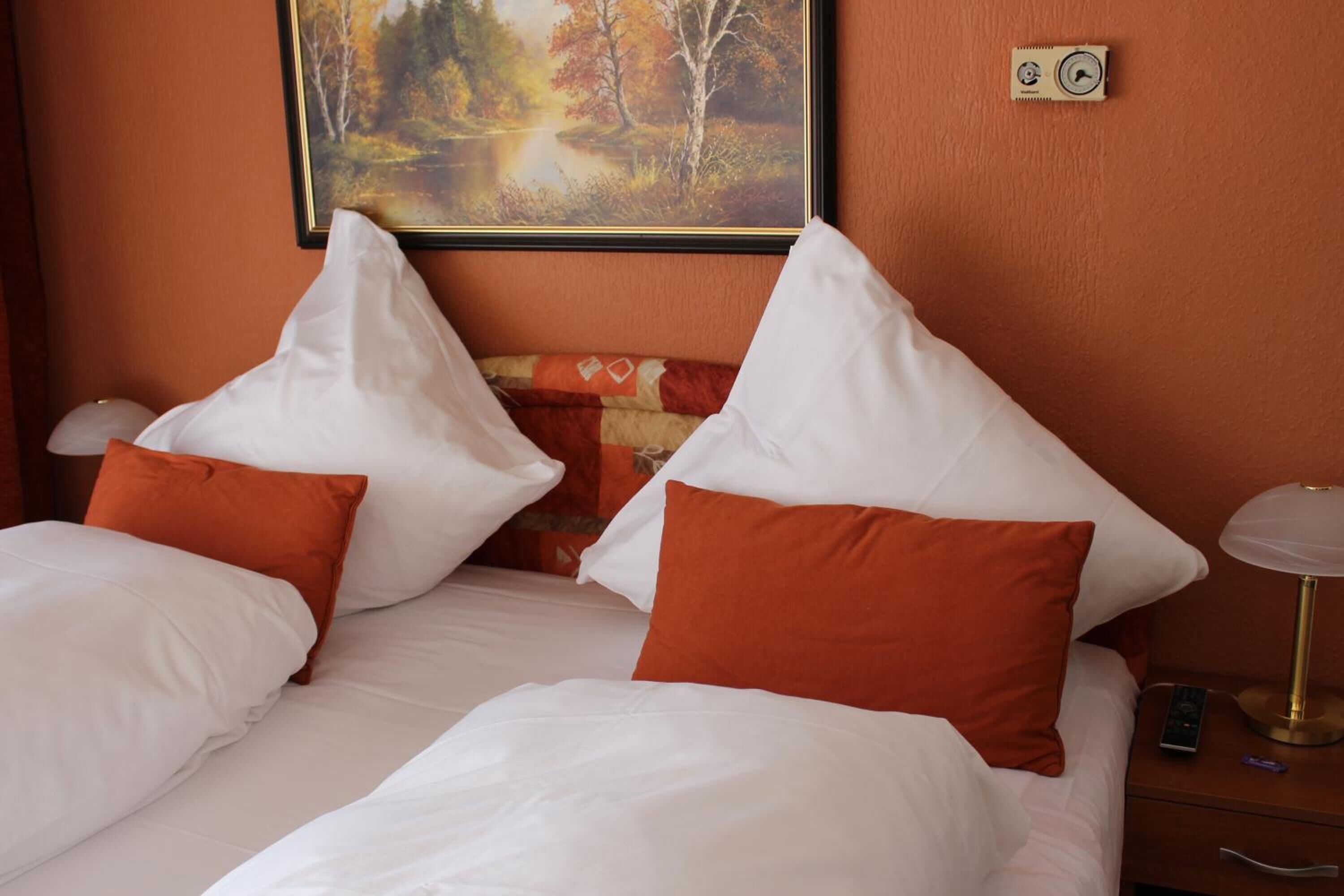 Una habitación de hotel con una cama, almohadas blancas y anaranjadas, un cuadro de un paisaje, una mesita de noche con una lámpara y un control remoto.