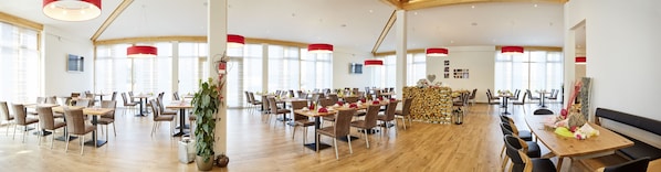 A spacious dining area with wooden tables and chairs, red pendant lights, and large windows allowing natural light.