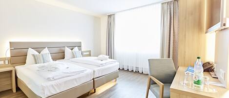 A hotel room with a double bed, bedside tables, a desk with a chair, and a large window with curtains.