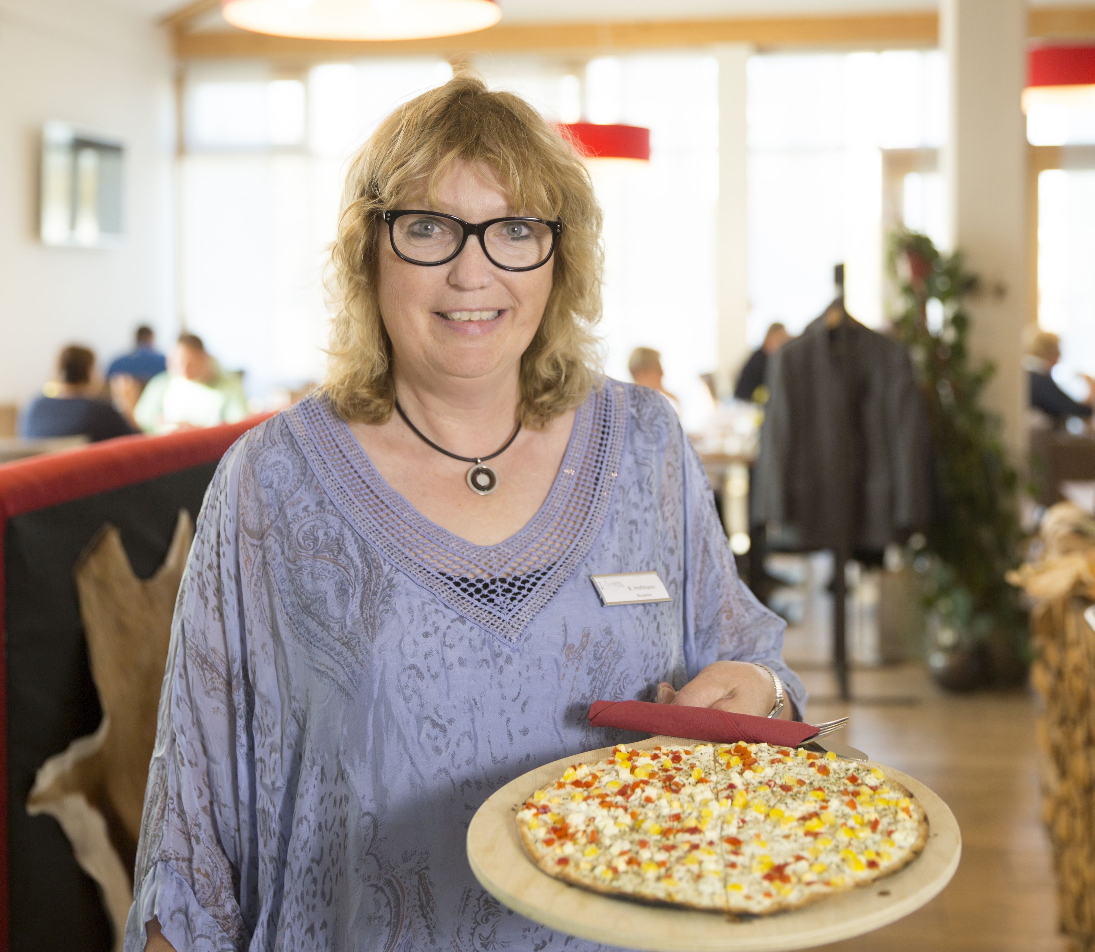 Una persona sosteniendo una pizza en una habitación con mesas y sillas, lo que sugiere un comedor.