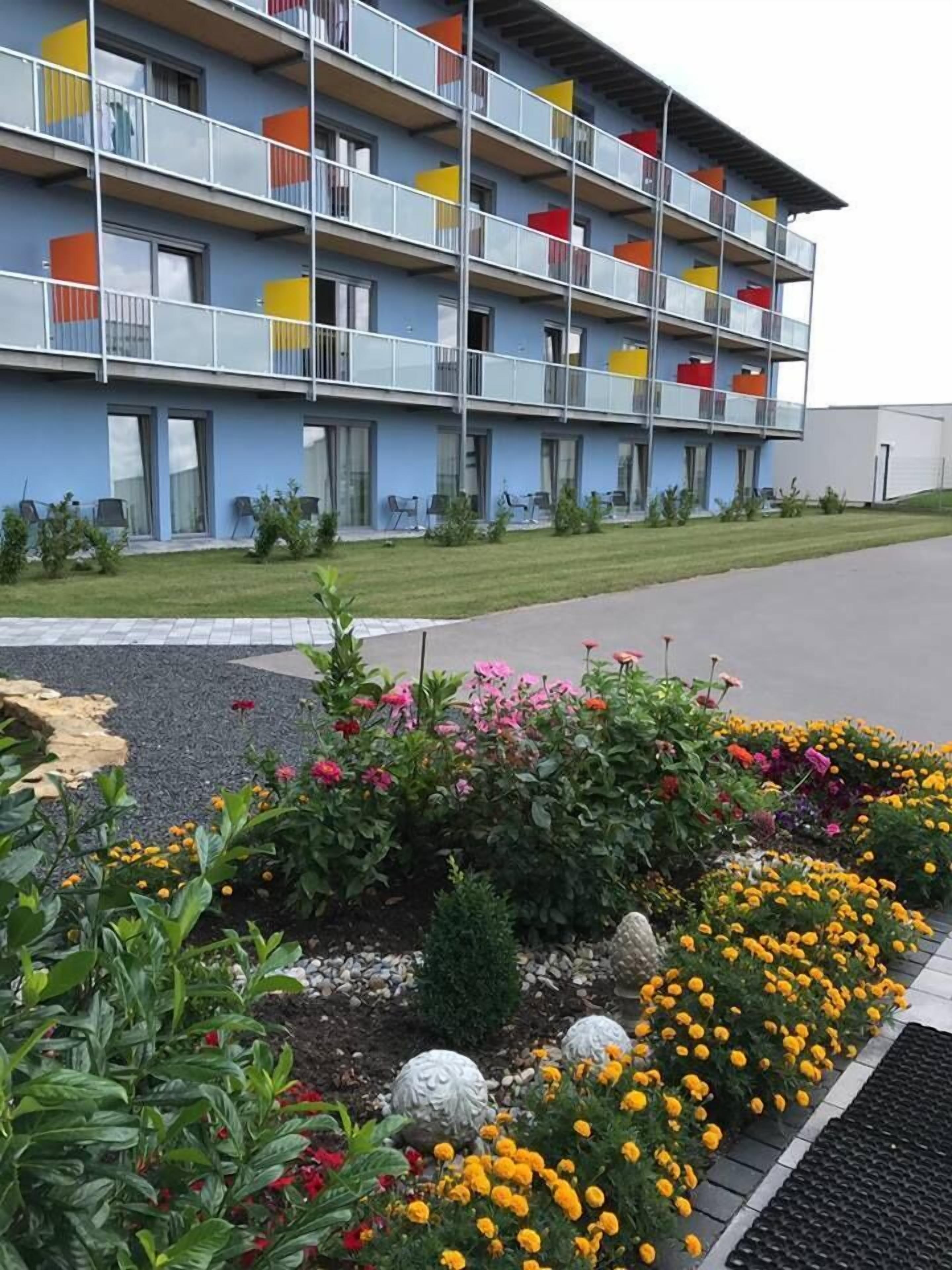 A multi-story hotel with balconies, a garden with various flowers, and a paved driveway.