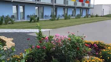 A multi-story hotel with balconies, a garden with various flowers, and a paved driveway.