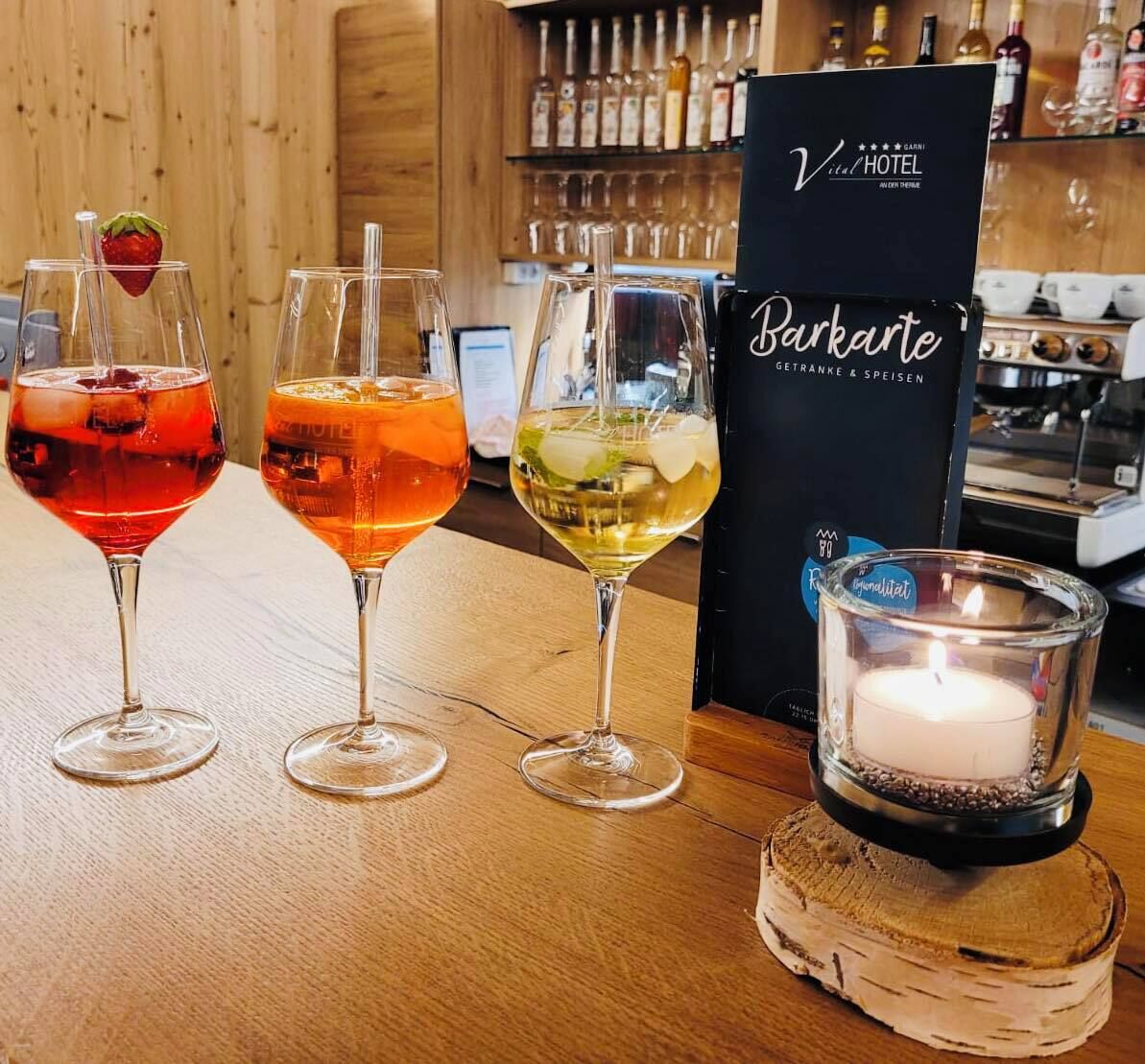 Trois verres contenant des boissons de couleurs différentes sont posés sur une table en bois. Une bougie se trouve dans un chandelier en verre, et on peut apercevoir, au fond, une enseigne indiquant « Vital Hotel ».