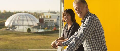 A man and woman standing on a balcony with a yellow wall, overlooking a park and a dome-shaped building.