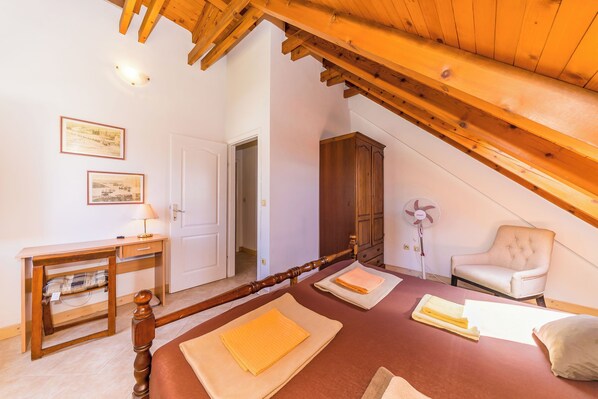 A bedroom with a wooden ceiling, a bed with beige and white bedding, a wooden desk with a lamp, a chair, and a wardrobe.