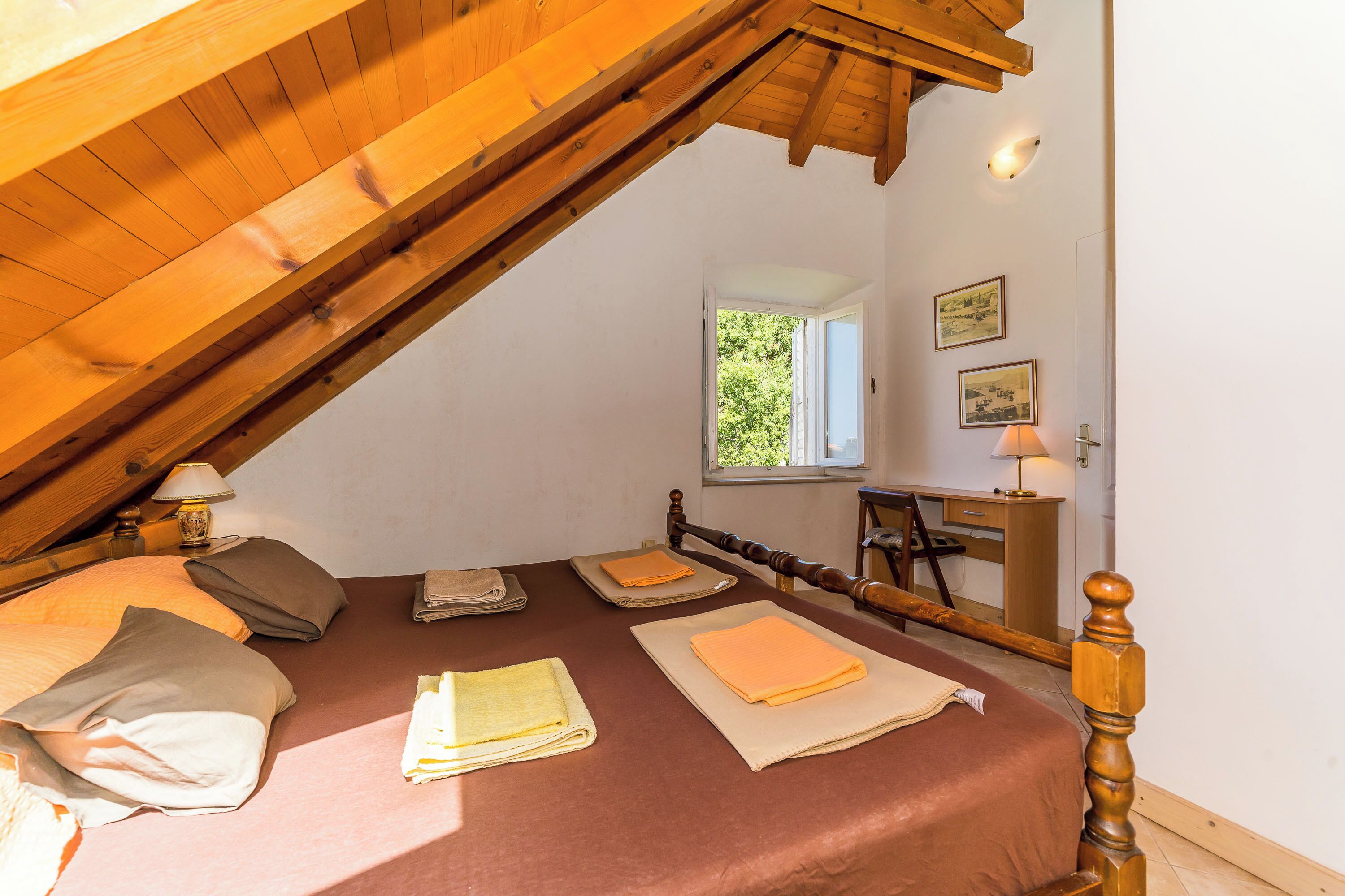 Un dormitorio acogedor en el ático, con techo inclinado, una cama con toallas ordenadas, una mesita de noche con lámpara, y una ventana con vistas a zonas verdes.