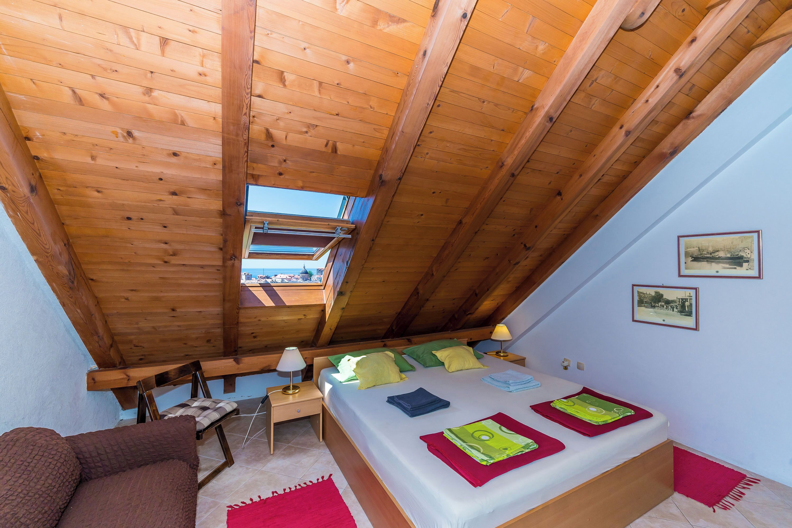 A bedroom with a sloped ceiling, a bed with pillows, a nightstand, a chair, and a window.