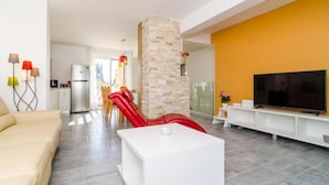 Una sala moderna con un sofá color beige, una mesa de centro blanca, una silla roja, un pilar de piedra, un televisor y una cocina al fondo.