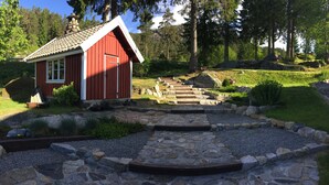 A red wooden cabin with a stone pathway leading to it, surrounded by a natural landscape with trees and rocks. - Hotell Eikerapen Gjestegard (Aseral)
