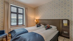 A double bed with a headboard, a bedside table with a lamp, a window with curtains, and a patterned wall. - Hotell Eikerapen Gjestegard (Aseral)