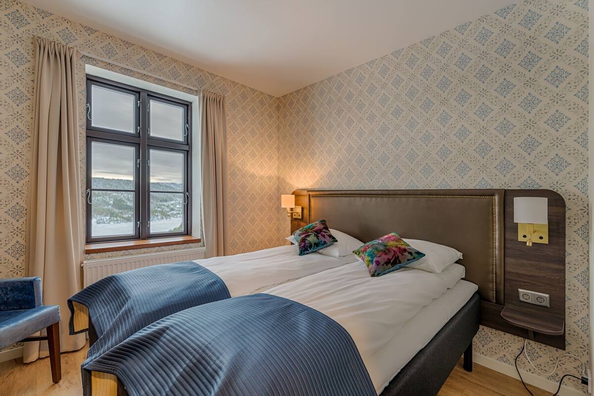 A double bed with a headboard, a bedside table with a lamp, a window with curtains, and a patterned wall.
