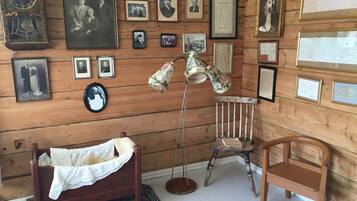 A room with wooden walls, a vintage clock, multiple framed photographs, a vintage lamp, a wooden chair, and a small wooden bed.