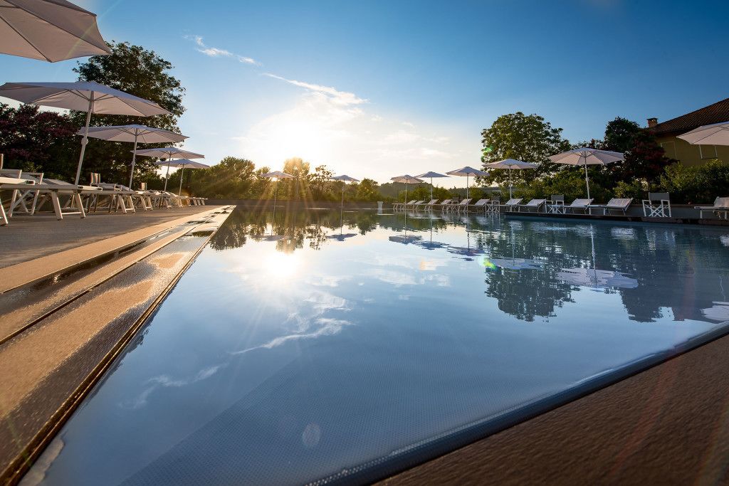 Un espace aménagé au bord de la piscine, avec des chaises longues et des parasols, où le soleil et les arbres se reflètent.