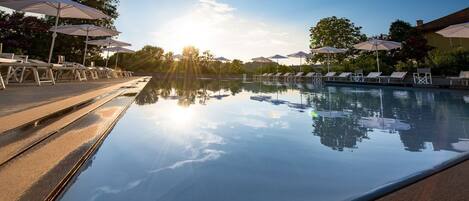 Un espace aménagé au bord de la piscine, avec des chaises longues et des parasols, où le soleil et les arbres se reflètent.