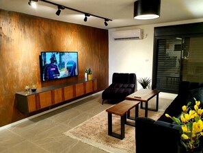 Una sala de estar moderna con televisor de pantalla plana, un sofá oscuro, una mesa de centro de madera y una planta en maceta.
