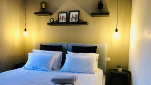 Una cama bien hecha con ropa de cama blanca y toallas enrolladas, una mesita de noche con una planta, y estantes en la pared con obras de arte enmarcadas y un reloj.