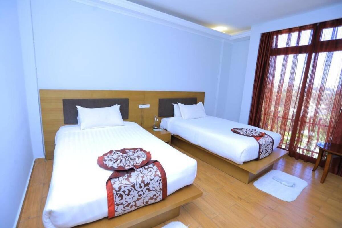 A hotel room with two beds, wooden headboards, white bedding, and a large window with curtains.
