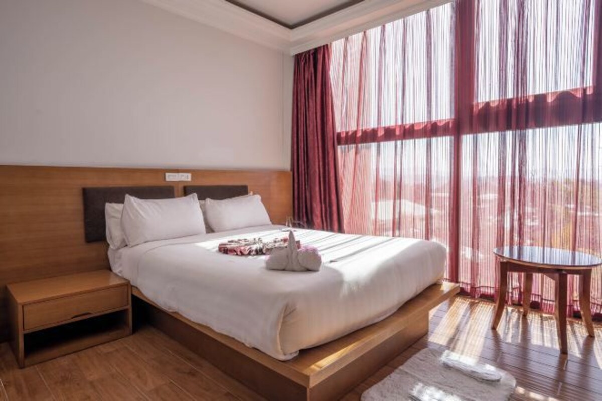 Una habitación de hotel con una cama de madera, una mesita de noche, una pequeña mesa y cortinas traslúcidas.