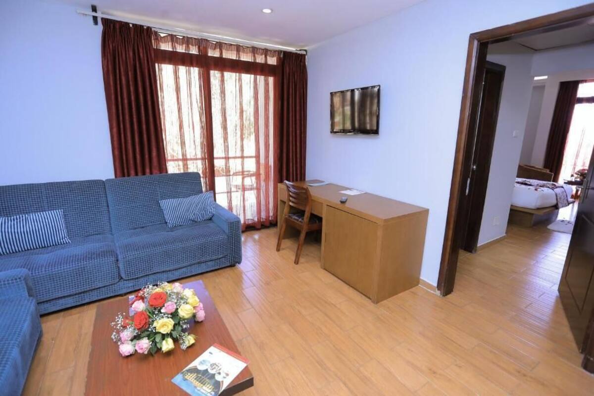 A living room with a sofa, a wooden coffee table with a book and flowers, a desk with a chair, and a TV on the wall.