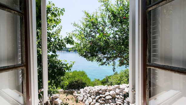 A view through an open window with a stone wall and greenery in the foreground, overlooking a body of water and distant land. - Guest House Simunovic (Dubrovnik)