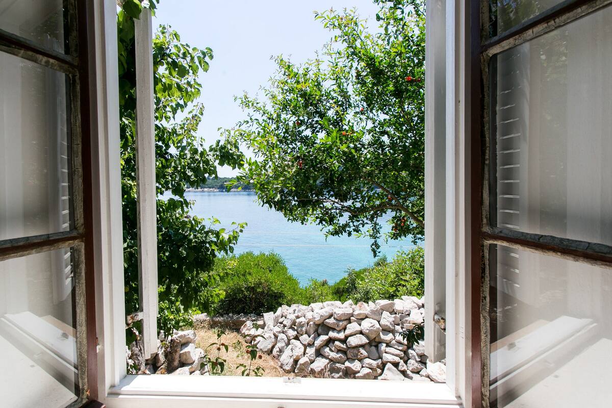 A view through an open window with a stone wall and greenery in the foreground, overlooking a body of water and distant land.